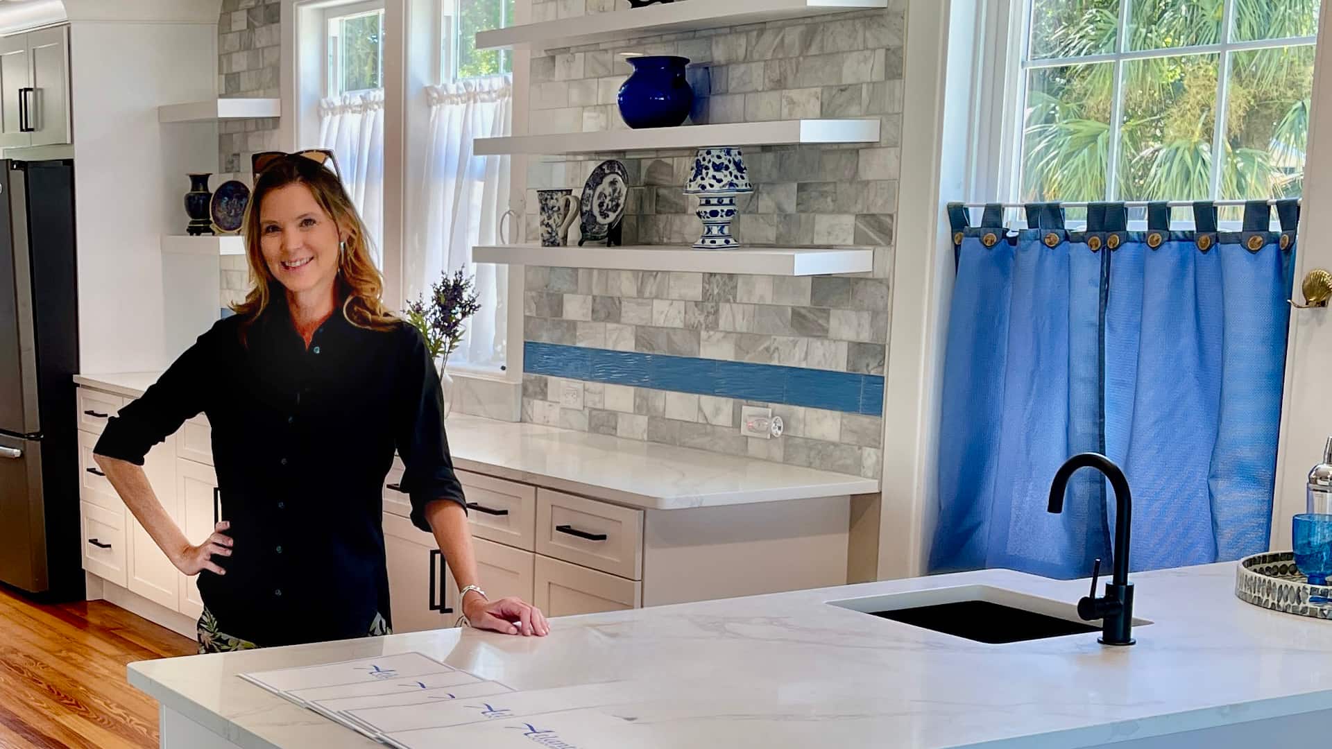 Kacie, lead designer at Timeless Kitchen & Bath Remodeling, standing in a remodeled Ormond Beach kitchen with white cabinetry, marble countertops, open shelving, and blue coastal accents.